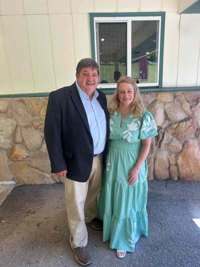 Man in a dark blazer and light shirt stands beside a woman in a turquoise floral maxi dress outside a stone‑and‑siding building with a window behind them.